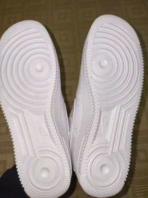 Nike Air Force 1 helvita sneakers - Nike Air Force 1 sneakers i helvitt med klassisk design, perforerad tå och platt sula. Skorna har snörning med metallbricka märkt 'AF1' och är tillverkade i skinn med textilfoder. Perfekt för dig som gillar stilrena och tidlösa sneakers.