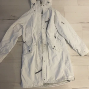 Vit skaljacka från Didriksons - Säljer en vit skaljacka från  Didriksons med huva och flera praktiska fickor med knappar. Jackan har lång ärm och dragkedja framtill, samt logotyp på ärmen. Perfekt för kalla och blöta dagar, riktigt clean och stilren look.