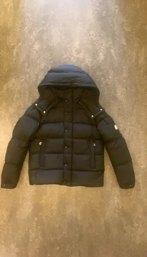 Svart Moncler vezere dunjacka med huva - Snygg svart dunjacka från Moncler med avtagbar huva, dragkedja och knappar framtill. Jackan har klassisk puffer-design, två fickor med dragkedja och logotyp på ärmen. Perfekt för kalla dagar och riktigt stilren look.