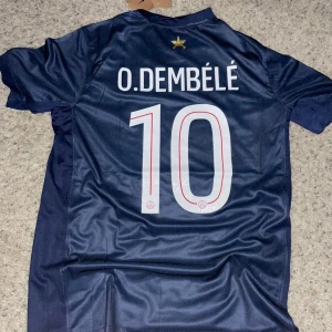 Blå PSG fotbollströja Dembele/Doué - Säljer en mörkblå PSG fotbollströja i polyester med tryckta namn och nummer: O. Dembélé #10 och D. Doué #14. Tröjan har korta ärmar, guldstjärna ovanför namnet och UEFA Foundation-märke på ärmen. Snygg design med vita och röda detaljer.