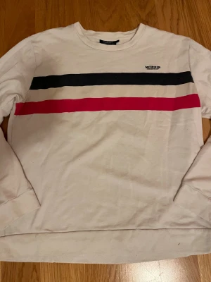 Vit tröja med ränder från Morris Company - Vit långärmad tröja från Morris Company i bomull. Framsidan har två breda horisontella ränder i blå och rött över bröstet samt en liten broderad logga. Tröjan har rund hals och klassisk passform, perfekt för en chill och stilren look. Kan gå ner i pris vid snabb och smidig affär, skriv vid frågor!