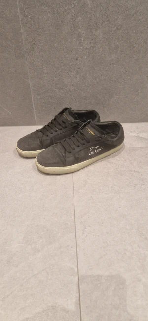 Svarta Saint Laurent sneakers - Svarta sneakers från Saint Laurent med vit text på sidan och guldfärgad logga på plösen. Skorna har snörning, rund tå och platt sula. Ovandelen är i canvas med detaljer i skinn, vilket ger en cool och urban vibe. Perfekt för dig som gillar stilrena och exklusiva sneakers. Vet ej om de e fejk eller inte.