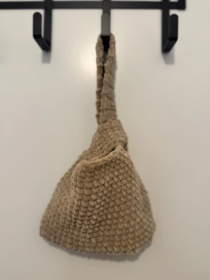 Beige virkad väska  - Supermysig beige liten pås- väska med grovstickad struktur och bred ren som går att hålla i handen  Väskan har en mjuk och fluffig känsla, perfekt för dig som gillar unika accessoarer. Den har en enkel, fyrkantig form men blir mer rund när du stänger väskan genom att trä in ena handtaget in i det andra. Perfekt att ha på språng 
