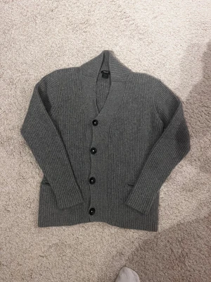 Grå ribbad kofta från Filippa K - Säljer en stilren grå kofta från Filippa K i storlek S. Koftan är ribbstickad med fyra svarta knappar framtill och har två fickor. Materialet är 73% bomull och 27% lammull, vilket gör den både mjuk och varm. Perfekt för lager-på-lager under kyliga dagar.