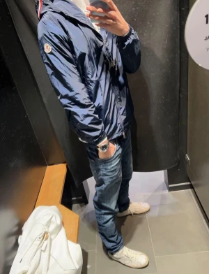 Mörkblå vindjacka från Moncler - Snygg mörkblå vindjacka från Moncler med klassisk logga på ärmen och dragkedja framtill. Jackan har huva, elastiska muddar och är tillverkad i glansig polyester. Perfekt för dig som vill ha en stilren och sportig look.