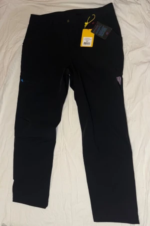 Svarta täckbyxor från Klättermusen XXL - Svarta täckbyxor från Klättermusen i storlek XXL. Byxorna har en rak passform och är tillverkade i slitstarkt syntetmaterial, perfekta för utomhusaktiviteter. Diskret Blåkläder-logga på benet och praktiska fickor. Snygg och funktionell design.