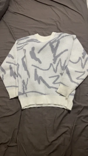 Vit och grå stickad tröja med mönster - Säljer en vit stickad tröja med grått abstrakt mönster. Tröjan har rund hals och ribbade muddar vid ärmslut och nederkant. Perfekt för dig som gillar unika prints och vill ha en mysig, stilren look.