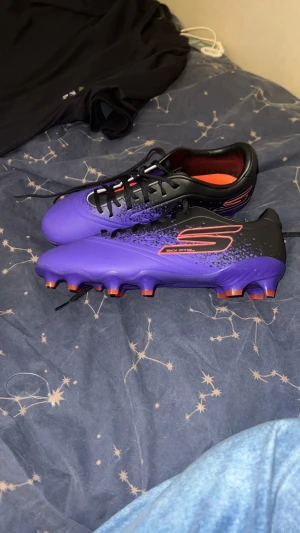 Lila och svarta fotbollsskor Skechers - Säljer ett par fotbollsskor från Skechers i en snygg lila och svart färg med orange detaljer. Skorna har snörning och en låg profil med platt sula och rund tå. Perfekt för dig som vill sticka ut på planen med cool design och bra grepp.