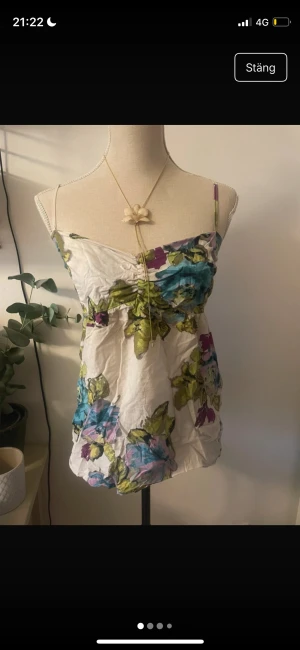 Blommig linnetopp med tunna axelband - Söt linnetopp med blommigt mönster i blått, grönt, lila och rosa på vit botten. Toppen har tunna axelband och lätt rynk vid bysten för en feminin look. Perfekt för varma dagar och ger en fräsch vibe till din outfit.