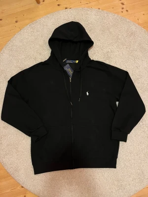 Svart hoodie från Polo Ralph Lauren - Svart hoodie från Polo Ralph Lauren i barnstorlek 2XL vilket motsvarar storlek M/L i vuxenstorlek. Tröjan har huva med snörning, dragkedja framtill och två fickor. Klassisk vit broderad logga på bröstet. Tillverkad i mjuk bomull, perfekt för chill dagar. Snygg och enkel design som funkar året runt. Lapp har vid ett tillfälle tidigare blivit avtagen men tröjan har aldrig varit använd. Pris kan diskuteras. 