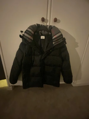 Svart pufferjacka från Burberry - Svart pufferjacka från Burberry med klassisk rutig krage i grått, svart, vitt och rött. Jackan har en rejäl dragkedja med Burberry-logga och en vadderad, boxig siluett. Perfekt för kalla dagar äkta pris kan diskuteras 