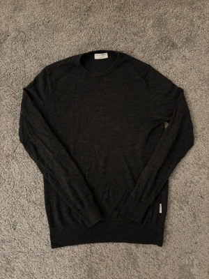 Mörkgrå merinoullströja Tiger of Sweden - Snygg mörkgrå stickad tröja från Tiger of Sweden i mjuk merinoull. Rund halsringning och långärmad modell med ribbade muddar. Perfekt för dig som gillar stilrena och klassiska plagg med skön passform. Plagget skickas inom 24 h efter köp! Hör av dig vid frågor…