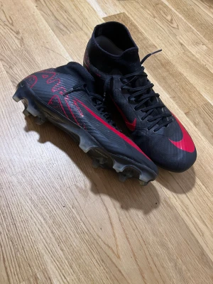 Nike Mercurial fotbollsskor svart/röd - Säljer ett par Nike Mercurial fotbollsskor i svart med röda detaljer och logga. Skorna har snörning, högre strumpliknande skaft och genomskinlig sula med fasta dobbar. Lätta och smidiga för snabba rörelser på planen. Perfekta för dig som vill sticka ut med stil.
