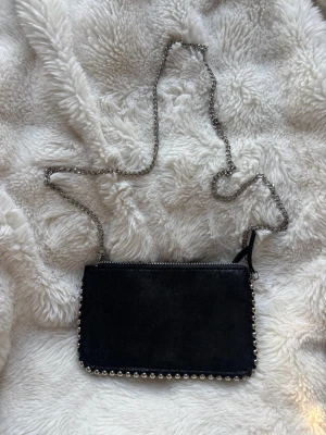 Svart axelväska från Zara med kedja och nitar - Snygg svart axelväska i mocka med silverfärgad kedja som axelrem och coola nitar längs kanterna. Väskan har dragkedja upptill och flera fack inuti för smidig organisering. Lika perfekt till vardag som till fest. Kommer med en kedja som man lätt kan ta av om man vill göra väskan till en kuvärtväska i stället. Köpt på Zara för ung 2 år sen. 
