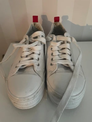 Vita sneakers från Guess med röd detalj - Snygga vita sneakers från Guess med klassisk låg modell, vita snören och silvriga öljetter. Sidan pryds av Guess logga i rött och skon har en röd hälflik. Ovandelen är i textil och sulan är vit med Guess-tryck runtom.