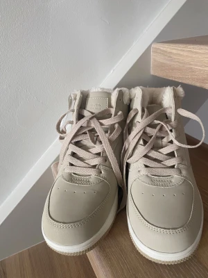 Beige sneakers från Lejon, storlek 37 - Snygga beige vinter sneakers från Lejon med högt skaft och mjukt foder. Skorna har snörning och en platt sula, perfekta för dig som gillar stilrena och bekväma skor. Utsidan är i syntetmaterial och insidan är fodrad med fluffigt tyg för extra komfort. Aldrig använt. 