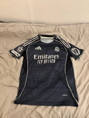 Real Madrid Champions League tröja - Säljer en marinblå Real Madrid Champions League fotbollströja från Adidas med vita detaljer och tunna ränder. Tröjan har korta ärmar, broderat klubbmärke, Adidas-logga och sponsortryck. Materialet är lätt och andas, perfekt för träning eller match. Får med tagen till tröjan.