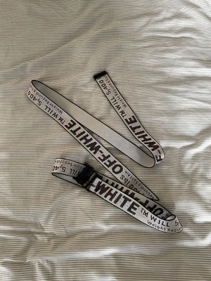 Vit Off-White bälte  - Säljer ett vitt industribälte från Off-White med svart och röd text längs hela bandet. Bältet har en robust svart spänne och är tillverkat i syntetmaterial. Perfekt för att ge outfiten en streetwear-vibe med tydlig logga och cool design.