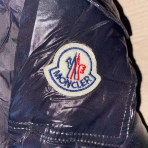 Svart Moncler pufferjacka med huva - Säljer en svart pufferjacka från Moncler med glansig finish och huva. Jackan har dragkedja framtill, två fickor med dragkedja och klassisk Moncler-logga på ärmen. Insidan har ett unikt serietidningsmärke. Perfekt för kalla dagar och riktigt snygg streetstil.