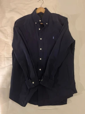 Mörkblå skjorta från Ralph Lauren - Klassisk mörkblå skjorta från Ralph Lauren med broderad logga på bröstet. Skjortan har långa ärmar, knappar framtill och button down-krage. Tillverkad i mjuk bomull som känns skön mot huden. Perfekt för en stilren och tidlös look.
