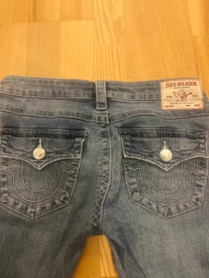 True Religion Joey bootcut jeans blå - Säljer ett par ljusblå True Religion Joey jeans med bootcut. Låg midja, klassiska fickor med lock och silverfärgade knappar. Snygga kontrastsömmar och ikonisk True Religion patch bak. Jeansen är i mjuk denim och har slitna detaljer vid benslut. W26