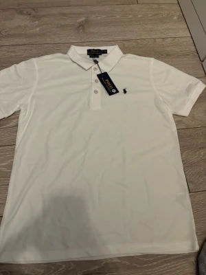 Vit pikétröja Polo Ralph Lauren M - Vit pikétröja från Polo Ralph Lauren i slim fit-modell. Klassisk krage, tre knappar framtill och den ikoniska broderade loggan på bröstet. Tillverkad i mjuk bomull som känns skön mot huden. Perfekt för en clean och stilren look.