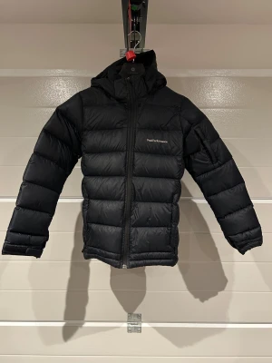 Svart dunjacka Peak Performance 160 - Svart dunjacka från Peak Performance i modellen JR Frost Down Jacket. Jackan har huva, dragkedja framtill och är quiltad med breda kanaler. Perfekt för kalla dagar och har en stilren look med diskret logga på bröstet.