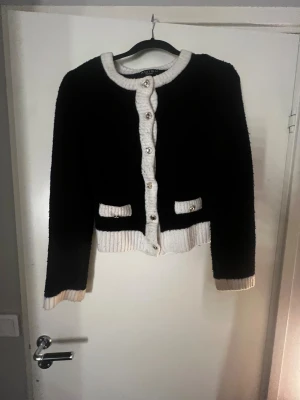 Svart och vit kofta från Amisu XS - Snygg svart kofta från Amisu i storlek XS med kontrasterande vita ribbade detaljer vid krage, knappslå, fickor och ärmslut. Koftan har silverfärgade knappar framtill och två dekorativa fickor. Perfekt för dig som gillar klassisk och trendig stil.