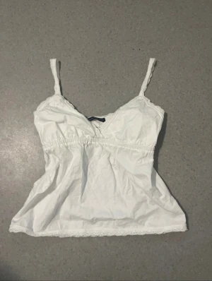 Vit linnetopp Brandy Melville - Supersöt vit linnetopp från Brandy Melville med tunna axelband och spetsdetaljer längs kanterna. Toppen har v-ringad hals.🩷