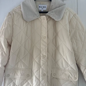 Quiltad jacka från NA-KD - Ljusgul/beige quiltad jacka ifrån NA-KD. Använd en gång förra året, så den är i nyskick. Pälsaktig krage högst upp, så den är perfekt nu inför vintern och även framåt våren!
