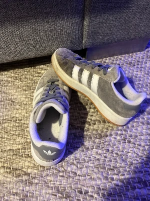 campus  - Säljer ett par Adidas Gazelle sneakers i grå mocka med klassiska vita ränder på sidorna och vit Adidas-logga på hälen. Skorna har snörning, platt gummisula i brunt och rund tå. Perfekt för dig som gillar retrostil och vill ha ett par snygga sneakers med ikonisk design.