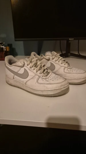 Nike Air Force 1 vita/ljusgråa sneakers - Nike Air Force 1 sneakers i klassisk vit färg med grå detaljer och swoosh. Lågt skaft, rund tå och platt sula. Skorna har snörning och är tillverkade i skinn med perforerade partier framtill för extra ventilation. Perfekt för dig som gillar streetstyle och tidlös design.