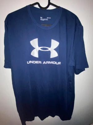 Blå Under Armour t-shirt med logga - Snygg marinblå t-shirt från Under Armour med stor vit logga och text på bröstet. Klassisk rund hals och korta ärmar. Tillverkad i mjuk bomull som känns skön mot huden. Perfekt för dig som gillar sportiga och stilrena plagg.