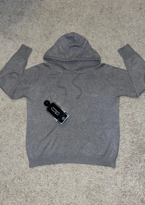 Grå Kashmir hoodie med snörning - En stilren grå hoodie med huva och snörning framtill. Tröjan har långärmade ärmar och ribbade muddar vid ärmslut och nederkant. Materialet känns mjukt och är bomull eller polyester, perfekt för chill dagar. Enkel design utan tryck eller mönster.