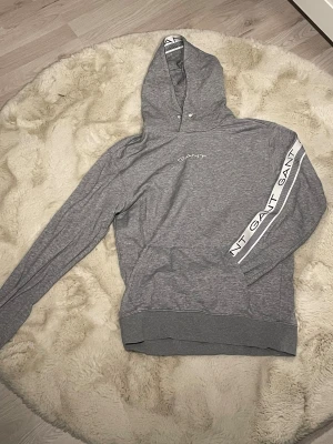Grå hoodie från GANT med logga - Snygg grå hoodie från GANT i storlek M. Tröjan har huva med vit kant och GANT-logga, samt en vit logga längs ena ärmen. Tillverkad i mjuk bomull och polyester, perfekt för chill dagar. Klassisk känguruficka framtill och ribbade muddar.