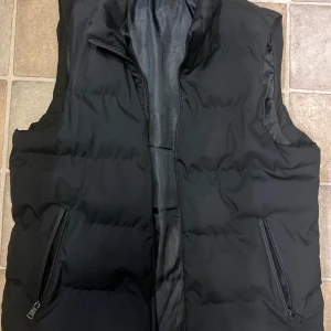 Svart dunväst med dragkedja, storlek M - Svart dunväst i storlek M med två praktiska fickor med dragkedja. Västen är quiltad och har en hög krage för extra värme. Tillverkad i 100% polyester, perfekt för lager-på-lager under kyliga dagar. Enkel och stilren design som funkar till många outfits.