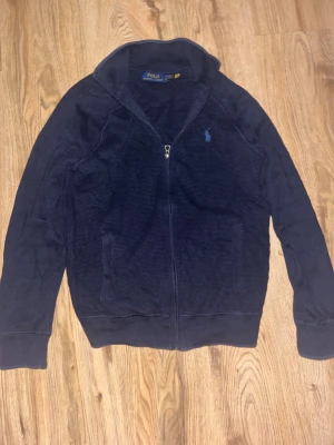 Ralph Lauren Zip  - Snygg marinblå kofta från Polo Ralph Lauren med dragkedja framtill och klassisk logga broderad på bröstet. Tröjan är stickad i bomull, har långärmad design och ribbade muddar vid ärmslut och krage. Perfekt för lager-på-lager under kyliga dagar.