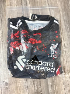 Liverpool x Adidas svart fotbollströja S - Snygg svart Liverpool fotbollströja från Adidas med röd och vit grafisk design och klubbmärke på bröstet. Tröjan har korta ärmar, vita axelränder och är tillverkad i lätt, ventilerande AEROREADY-material. Perfekt för dig som vill sticka ut på planen.