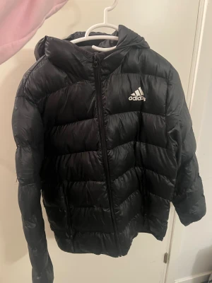 Svart pufferjacka från Adidas Strl 164 men passar S/M - Svart pufferjacka från Adidas med huva och dragkedja framtill. Jackan har Adidas-logga på bröstet och är vadderad för extra värme. Perfekt för kyliga dagar och har en sportig vibe. Passar dig som gillar streetwear och vill hålla dig varm.