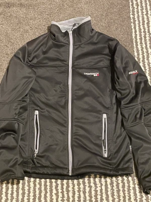 Svart softshelljacka från True North - Svart softshelljacka från True North med grå detaljer och hel dragkedja framtill. Jackan har två fickor med dragkedja och logga på bröstet samt ärmen. Insidan är fodrad med mjuk grå fleece för extra komfort. Perfekt för kyliga dagar.