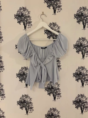 Ljusblå blus med puffärmar från Gina Tricot - Supersöt ljusblå blus från Gina Tricot med puffiga korta ärmar och bred urringning. Blusen har en knytning framtill och är tillverkad i ett mjukt, lätt polyester-tyg som faller snyggt. Perfekt för dig som gillar romantiska och trendiga detaljer.