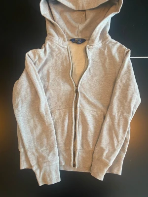 Grå hoodie från Polo Ralph Lauren - Säljer en grå hoodie från Polo Ralph Lauren med dragkedja framtill och huva. Tröjan har långa ärmar, två fickor och är tillverkad i mjuk bomull. Perfekt för chill dagar och enkel att matcha med jeans eller joggers.