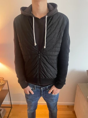 Svart cardigan jacka från J.Lindeberg - Svart dunväst från J.Lindeberg i storlek S. Framsidan är quiltad med dragkedja och har stickade partier på ärmar och rygg. Snygg, stilren design med diskret ficka på ärmen och ribbade muddar. Perfekt för lager-på-lager under kyliga dagar. Kan gå ner i pris vid snabb affär 