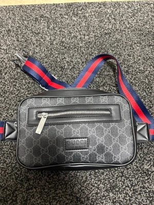 Gucci magväska med GG-mönster - Snygg magväska från Gucci i svart med klassiskt GG-mönster och silverfärgad dragkedja. Väskan har en justerbar axelrem i blått och rött samt en praktisk ytterficka. Perfekt för att bära det viktigaste med stil.