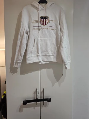 Vit hoodie från GANT med tryck - Säljer en vit hoodie från GANT med huva och känguruficka. Framtill finns ett stort tryck med GANT-logga, amerikansk sköld och texten 'American Sportswear' i guld och rött. Klassisk passform och ribbade muddar. Perfekt för en avslappnad och sportig stil.