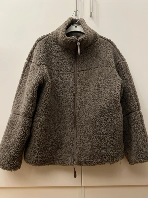 Beige/grå teddyjacka med dragkedja - Mysig mörkgrå teddyjacka med hög krage och hel dragkedja framtill från Gina tricot. Jackan har en boxig passform och är tillverkad i fluffigt teddy-material som håller dig varm. Perfekt för kyliga dagar och enkel att styla till olika outfits.