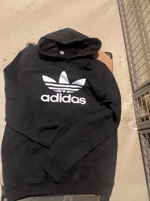 Svart Adidas hoodie med stor logga - Svart hoodie från Adidas med stor vit Trefoil-logga och text på bröstet. Klassisk modell med huva och ribbade muddar. Perfekt för en sportig och avslappnad stil. Tillverkad i mjukt material för extra komfort.