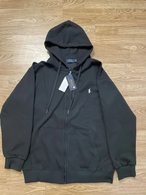 Svart hoodie från Polo Ralph Lauren - Svart hoodie med dragkedja från Polo Ralph Lauren. Klassisk design med huva, dragsnören och två fickor framtill. Liten vit broderad logga på bröstet. Tillverkad i mjuk bomullsblandning för en skön känsla. Passar M