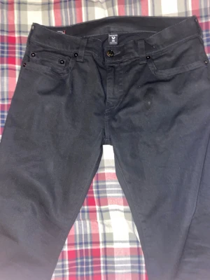 Svarta jeans från true religion  - Säljer ett par svarta jeans från true religion med klassisk femficksmodell och raka ben. Jeansen har svarta nitar och knappdetaljer samt diskret logga i linningen. Materialet är mjukt och stretchigt för extra komfort.