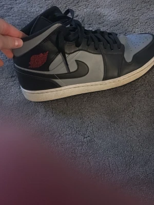 Air Jordan 1 Mid svart/grå - Snygga Air Jordan 1 Mid sneakers i svart och grått läder med klassisk röd Air Jordan-logga på sidan. Skorna har svart swoosh, svarta skosnören och vit sula. Perfekta för dig som gillar streetwear och vill ha en ikonisk look.